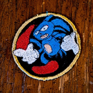May include: A circular embroidered patch featuring a blue character with white gloves and red shoes centred on a black background. The design is enclosed by a thin gold border, showcasing detailed stitching of the character's features and limbs.