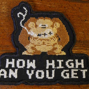 Puede incluir: Un parche bordado negro con un personaje de dibujos animados fumando una pipa y el texto "How high can you get?"