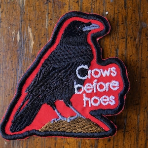 以下が含まれることがあります： 赤い縁取りと背景を持つ黒いカラスが描かれた刺繍パッチ。「Crows before hoes」というテキストが白い文字で含まれています。パッチは木製の表面に配置されています。