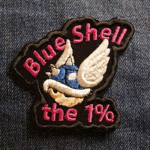 Op de afbeelding: Zwarte geborduurde patch met een blauwe schelp met witte vleugels en de tekst "Blue Shell the 1%" in roze.