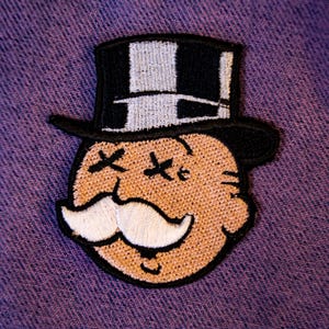 May include: An embroidered patch featuring a cartoon character with a tan face, a large white mustache, and black X-shaped eyes. The character wears a black and white top hat with a checkered pattern. The patch has a thick black border outline.