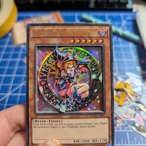 Puede incluir: Una carta de Yu-Gi-Oh! Dark Magician Girl con un diseño holográfico. La carta presenta una ilustración detallada del personaje, con texto en alemán. El ATK de la carta es 2000 y la DEF es 1700.