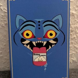 以下が含まれることがあります： 青いノート。猫の顔をデザイン。猫は黄色い目、赤い瞳、ピンクの舌。ノートには舌に「Notes and more」の文字。表紙は金の縁取り。
