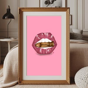 May include: Framed art print featuring a pop art style image of a mouth with glossy, pink lips and white teeth, holding a gold bullet. The background is a solid pink color. The frame is gold.