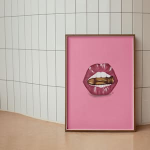 May include: A framed art print featuring a pink background with an illustration of a pair of lips with a bullet in the mouth. The lips are a deep pink color, and the bullet is gold. The frame is a dark brown color.