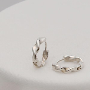 Puede incluir: Un par de pendientes de aro de plata con un diseño trenzado. Los pendientes son pequeños y delicados.