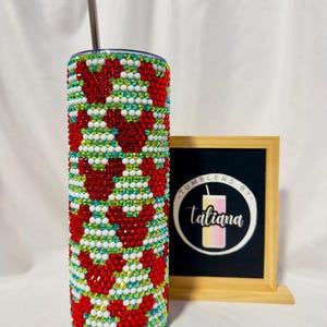 May include: A bedazzled tumbler with a red heart pattern, white, and blue accents. The stainless steel tumbler includes a silver straw. A framed sign with the text "Tumblers by Tatiana" is in the background.