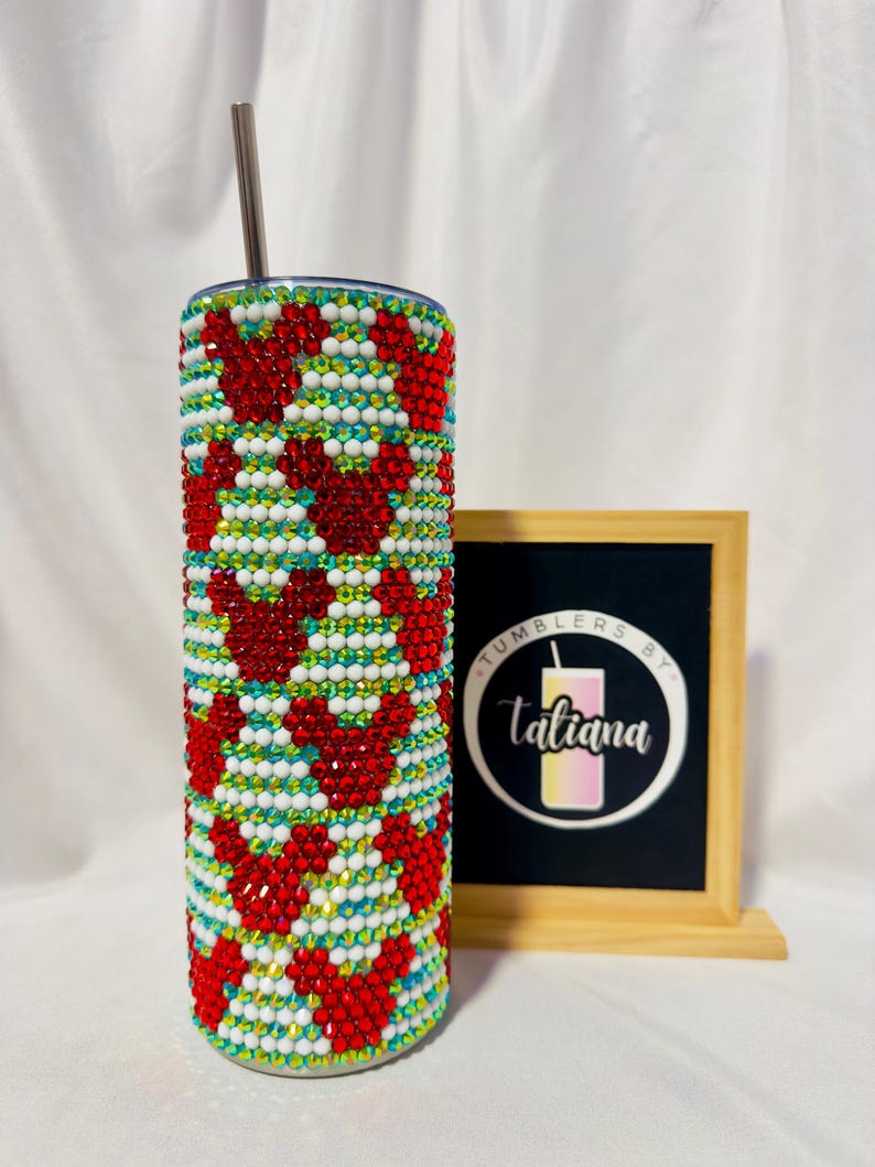 May include: A bedazzled tumbler with a red heart pattern, white, and blue accents. The stainless steel tumbler includes a silver straw. A framed sign with the text "Tumblers by Tatiana" is in the background.