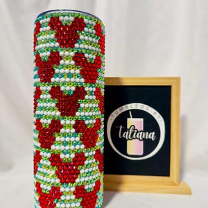 May include: A bedazzled tumbler with a pattern of red heart shapes, white, and iridescent rhinestones. The tumbler has a silver lid and is decorated with a variety of colorful gems. The tumbler is next to a framed sign with the text "Tumblers by Tatiana".