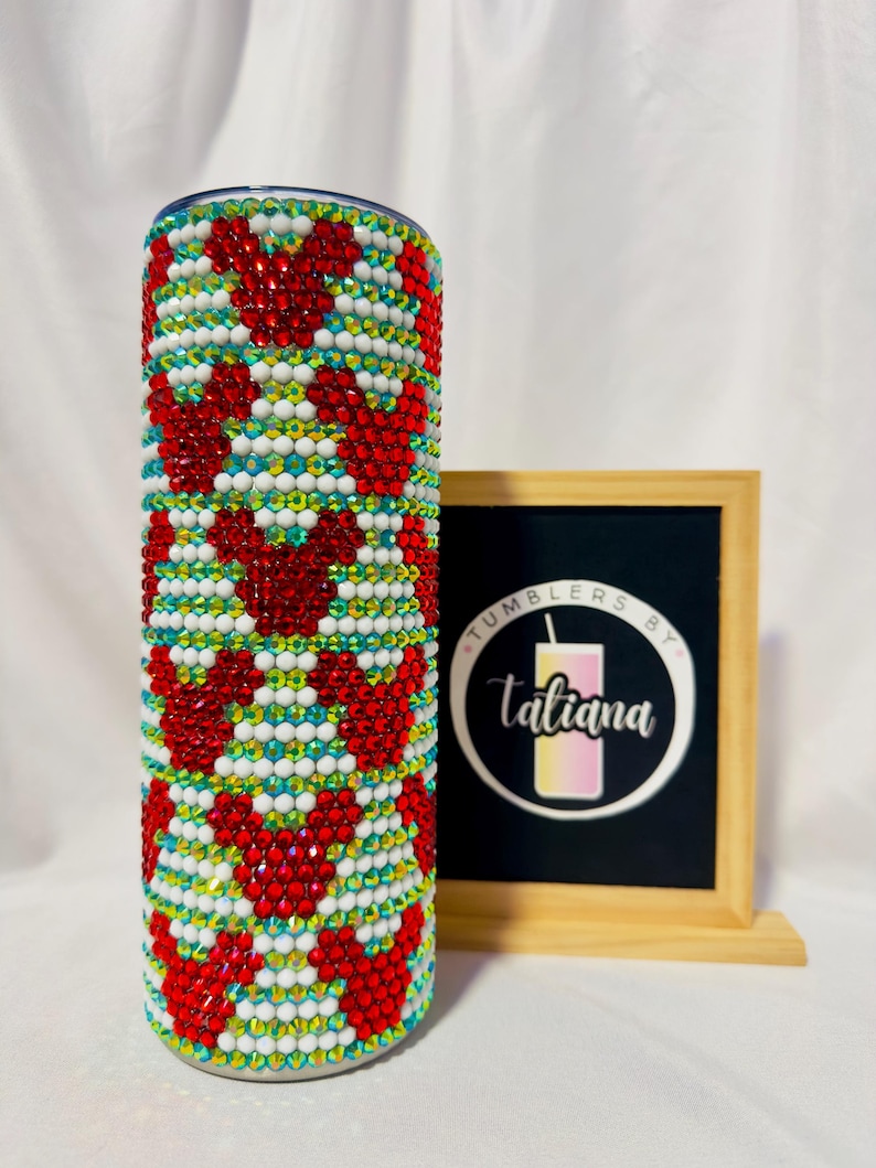 May include: A bedazzled tumbler with a pattern of red heart shapes, white, and iridescent rhinestones. The tumbler has a silver lid and is decorated with a variety of colorful gems. The tumbler is next to a framed sign with the text "Tumblers by Tatiana".