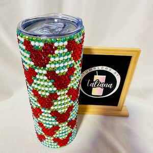 May include: A bedazzled tumbler with a clear lid. The tumbler is covered in red, white, and green rhinestones arranged in a chevron pattern. A small chalkboard sign with the text "Tumblers by Taliana" is to the right of the tumbler.
