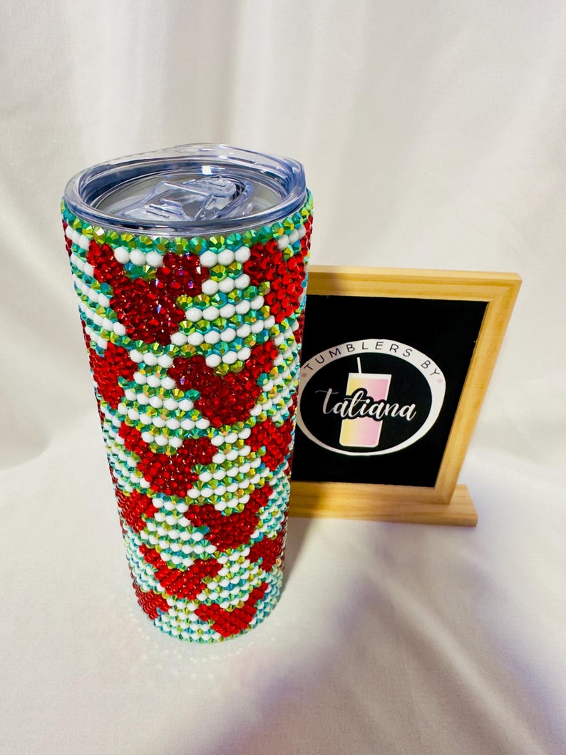 May include: A bedazzled tumbler with a clear lid. The tumbler is covered in red, white, and green rhinestones arranged in a chevron pattern. A small chalkboard sign with the text "Tumblers by Taliana" is to the right of the tumbler.