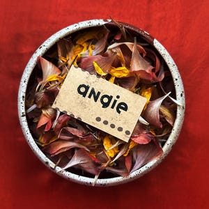 May include: A ceramic bowl filled with colorful flower petals, including yellow, orange, and red hues. A small, rectangular tag with the name "angie" printed in black letters is placed on top of the petals. The background is a solid red.