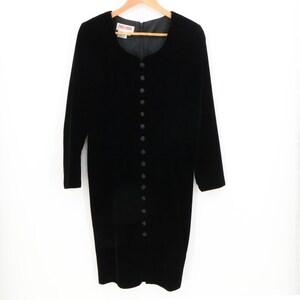 May include: A black velvet dress with long sleeves and a row of buttons down the front. The dress has a round neckline and is hanging on a wooden hanger against a white background. The dress appears to be knee-length.