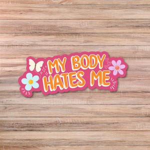 May include: A die-cut sticker with a pink background featuring the orange text MY BODY HATES ME. The design includes a white butterfly, a light blue flower, a purple flower, and small heart and star accents surrounding the bold lettering.