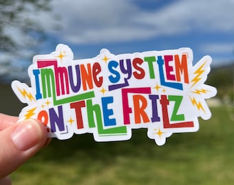 Pode incluir: Um autocolante colorido com a frase "Immune System On The Fritz" numa fonte multicolorida. O design inclui detalhes de estrelas e raios sobre um fundo branco.