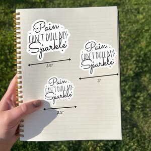 May include: Three white stickers with the text "Pain Can't Dull My Sparkle" in a cursive font. The largest sticker measures 3.5 inches, the middle one 3 inches, and the smallest 2.5 inches. The stickers are on a spiral notebook.
