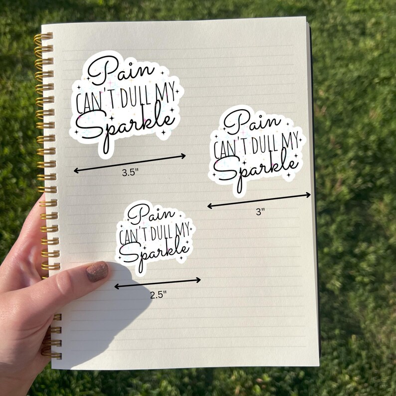 May include: Three white stickers with the text "Pain Can't Dull My Sparkle" in a cursive font. The largest sticker measures 3.5 inches, the middle one 3 inches, and the smallest 2.5 inches. The stickers are on a spiral notebook.