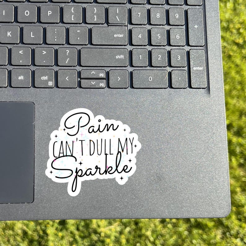 May include: A sticker with the text "Pain CAN'T DULL MY Sparkle" in a cursive font, with a black outline and white fill. The sticker is on a gray laptop keyboard.