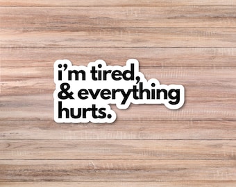 May include: A white sticker with black text that reads "I'm tired, & everything hurts." The sticker is rectangular with rounded corners. The background is a light brown wooden surface.