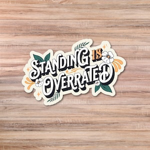 May include: A sticker with the phrase "STANDING IS OVERRATED" in a whimsical font. The design includes floral and leaf accents in green, white, and orange against a cream background. The sticker is set against a wooden background.