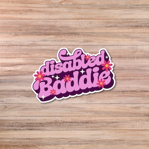 May include: A sticker featuring the words "disabled baddie" in a retro font, with a white and dark purple outline. Pink flowers and white stars are incorporated into the design. The sticker is placed on a wooden surface.