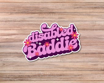 May include: A sticker with the words "disabled baddie" in a retro font, outlined in white and dark purple. Pink flowers and white stars decorate the design. The sticker is on a wooden surface.