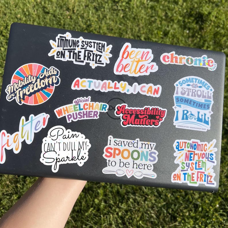May include: A black laptop covered in colorful stickers with various phrases, including "Mobility Aids are Freedom," "Actually I Can," and "I saved my SPOONS to be here." The stickers have a variety of fonts and designs.