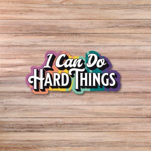 May include: A colorful sticker with the phrase "I Can Do Hard Things" in a bold, stylized font. The text is outlined in white and set against a backdrop of vibrant colors, including purple, orange, yellow, and teal. The sticker is placed on a wooden surface.