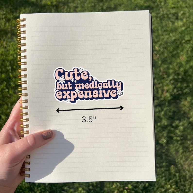 May include: A sticker with the text "Cute, but medically expensive" in pink and blue, with small white flower accents. The sticker is 8.89 cm long and is on a lined notebook.