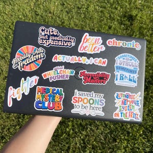 May include: A black laptop adorned with multiple vibrant stickers. The stickers display text such as "Accessibility Matters," "Been Better," and "Actually, I Can." The laptop is positioned against a background of green grass, with the stickers' messages clearly visible.