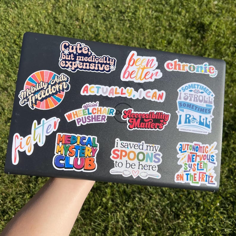 May include: A black laptop adorned with multiple vibrant stickers. The stickers display text such as "Accessibility Matters," "Been Better," and "Actually, I Can." The laptop is positioned against a background of green grass, with the stickers' messages clearly visible.