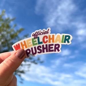 May include: A colorful sticker with the text "Official Wheelchair Pusher" in a rainbow of colors. The sticker is held against a backdrop of a blue sky with some clouds.