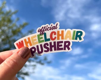 May include: A colorful sticker with the text "Official Wheelchair Pusher" in a rainbow of colors. The sticker is held against a backdrop of a blue sky with some clouds.