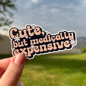 May include: A sticker with the phrase "Cute, but medically expensive" in a retro font. The text is in peach and white, set against a navy blue background, with floral and star accents. The sticker is held by a hand.