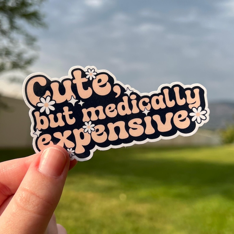 May include: A sticker with the phrase "Cute, but medically expensive" in a retro font. The text is in peach and white, set against a navy blue background, with floral and star accents. The sticker is held by a hand.
