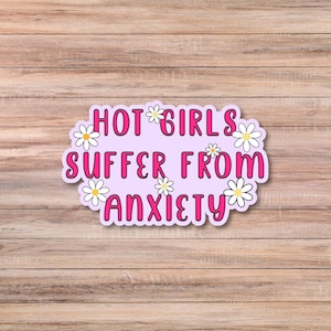 May include: A sticker with the text "HOT GIRLS SUFFER FROM ANXIETY" in pink lettering, surrounded by white daisy flowers, on a light purple background. The sticker is set against a wooden background.
