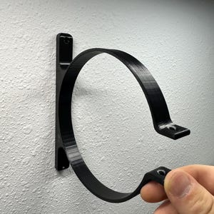 May include: A black, circular wall-mounted bracket, designed to hold an object. The bracket is made of a solid material and has a curved design with a flat mounting surface. The bracket is attached to a textured white wall.