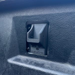 May include: A close-up of a black plastic component with the text "DIGIFAB" printed on it. The component has a unique shape with a recessed area and is mounted on a textured surface.
