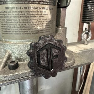 May include: A black, star-shaped knob with a textured surface and the letters "OF" embossed in the center. The knob is attached to a gray, industrial hydraulic shop press featuring a metal label with "IMPORTANT - BLEEDING INSTRUCTIONS" text.