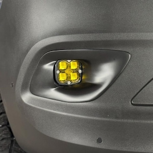 May include: Close-up of a vehicle's front bumper with a black housing containing four bright yellow LED lights. The bumper is a matte gray color, and the lights are square-shaped.