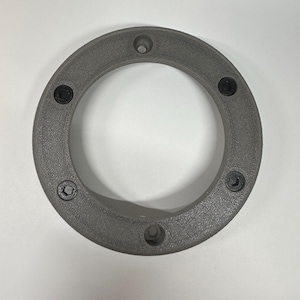 May include: A gray, circular object with a central opening, featuring evenly spaced holes and black screws. The object appears to be made of a textured material, possibly plastic or a composite. The design suggests a mechanical or structural component.