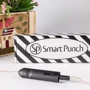 May include: A black Smart Punch tool with a needle and thread, next to a black and white striped box with the words "Smart Punch" on it. A wooden planter box with flowers and a decorative needlepoint design is in the background.
