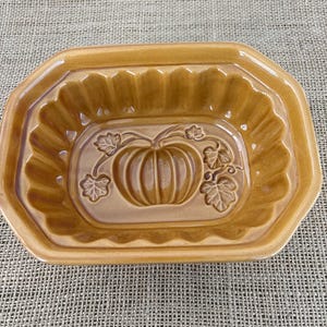 May include: A ceramic, rectangular baking dish in a warm, golden-brown hue. The center features a raised pumpkin design with leaves. The dish has a scalloped edge and is ideal for fall-themed baking and serving.