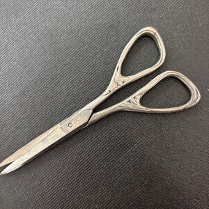 May include: Silver-toned scissors with ornate handle detailing. The scissors have a pointed tip and are designed for precision cutting. The handles feature a decorative floral pattern. The scissors are likely vintage or antique.
