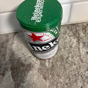 May include: A green Heineken Experience tin with the Heineken logo and text. The tin features a red star and the words "Amsterdam 1873". The top of the tin has water droplets.