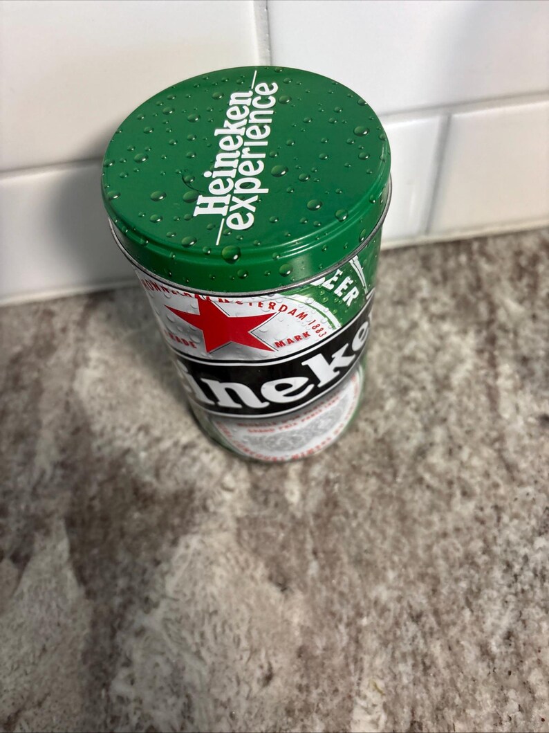 May include: A green Heineken Experience tin with the Heineken logo and text. The tin features a red star and the words "Amsterdam 1873". The top of the tin has water droplets.