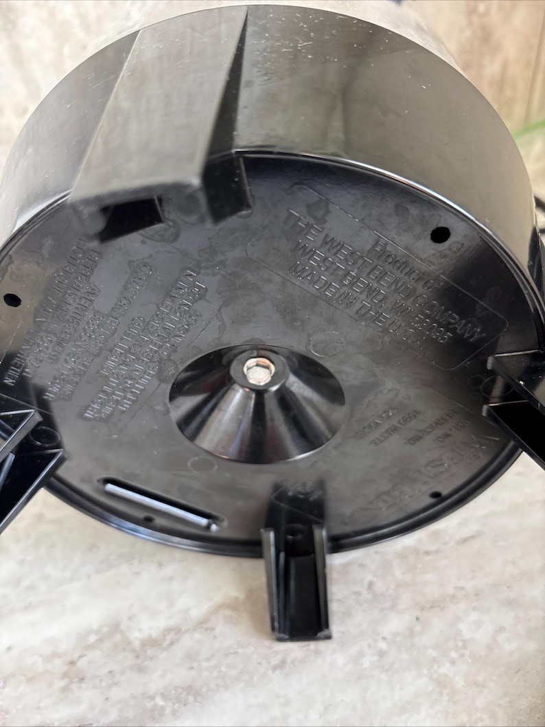 Puede incluir: Base de electrodom&eacute;stico de pl&aacute;stico negro con tres patas y un tornillo central. La base tiene el texto "THE WEST BEND COMPANY" y "MADE IN THE USA" impreso. La base del electrodom&eacute;stico est&aacute; sobre una superficie clara.