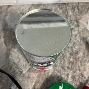 May include: A silver metal can with green and white branding. The cylindrical can has a flat top and a red accent at the base. The can is placed on a speckled surface.