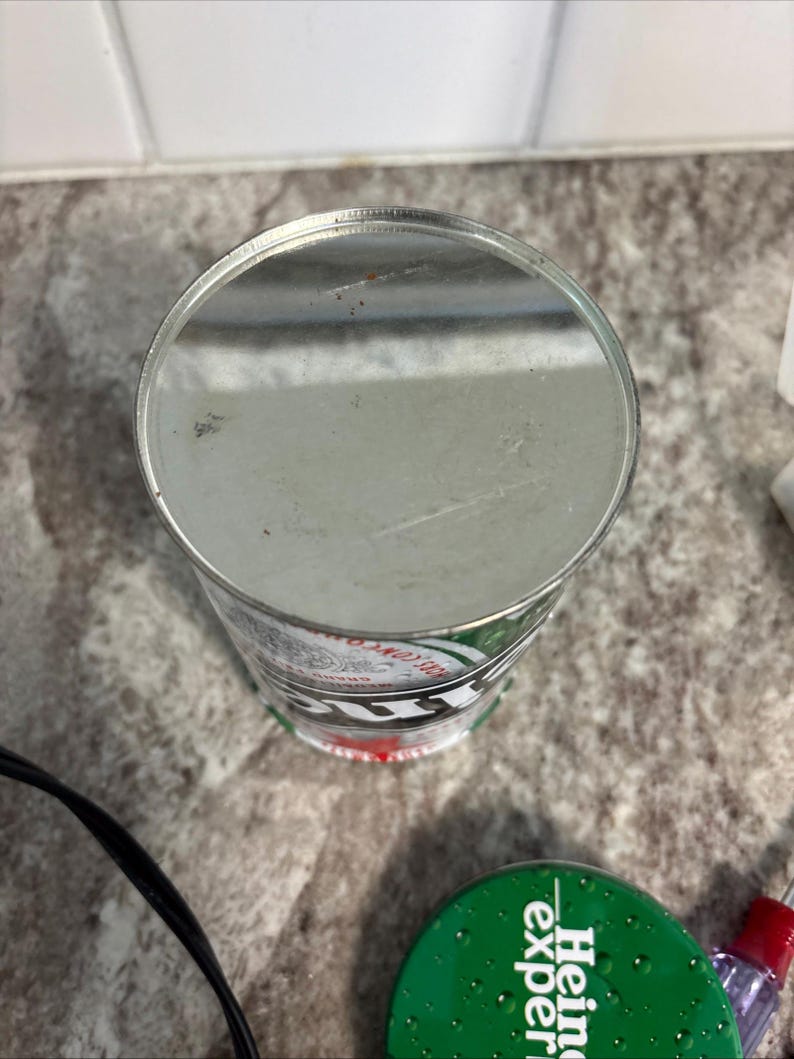 May include: A silver metal can with green and white branding. The cylindrical can has a flat top and a red accent at the base. The can is placed on a speckled surface.